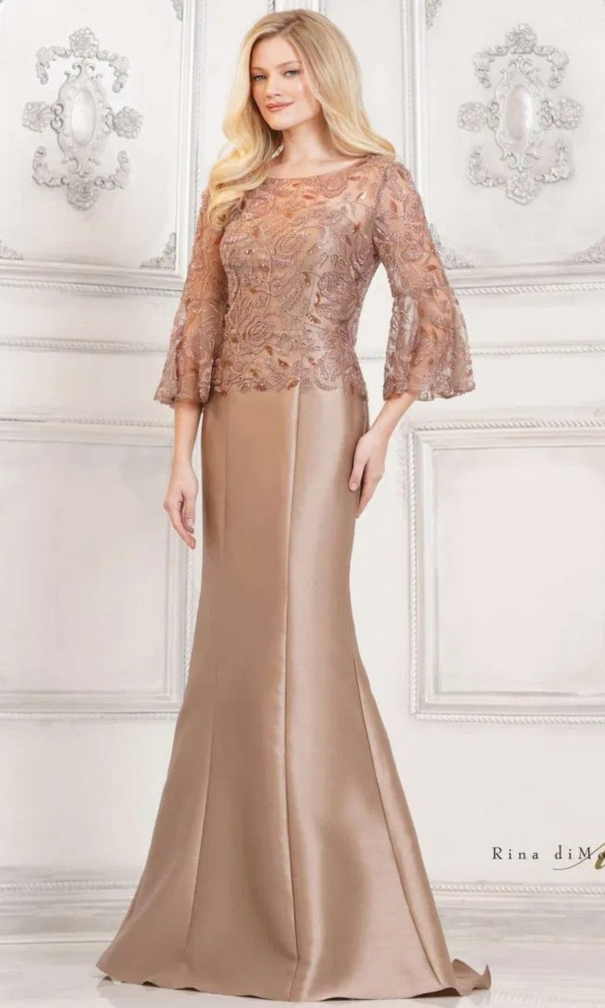 A woman wears the Rina Di Montella RD2939 long formal dress in taupe, featuring a mermaid skirt, lace sleeves, and a fitted satin bodice, standing before an ornate light-colored wall.